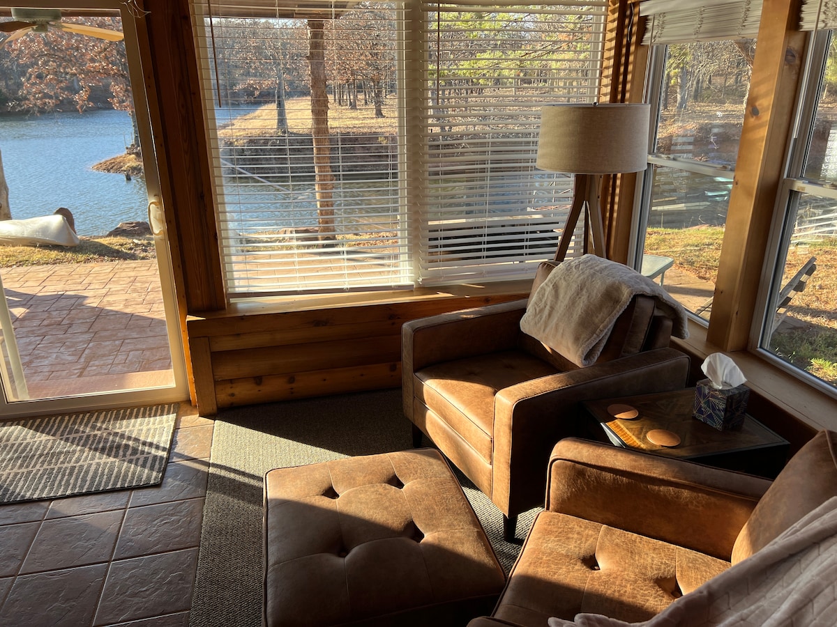 A sunlit sitting area features two brown leather armchairs beside a small table. A lamp provides additional light, while large windows offer a view of the tranquil pond and surrounding nature. The flooring consists of large, warm-toned tiles.