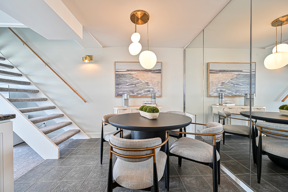 A dining area is highlighted by a round table surrounded by four upholstered chairs. A contemporary light fixture hangs overhead, while a large mirror reflects the room's design. The staircase is visible in the background, leading to the upper level.
