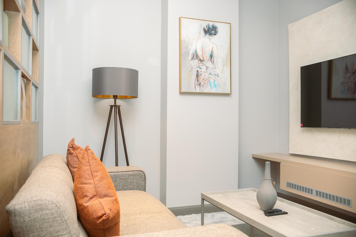 A comfortable seating area is featured, with a light-colored sofa accented by an orange decorative pillow. A stylish floor lamp stands beside the sofa, complementing a wall-mounted television. An abstract artwork adds character to the room, enhancing the modern design elements.