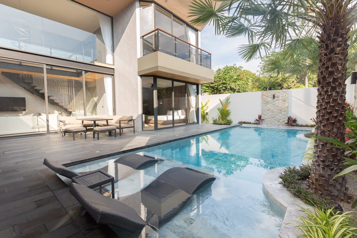 The outdoor area features a private saltwater swimming pool, surrounded by lush greenery and tropical plants. Two sun loungers are positioned partially submerged in the shallow end, while a covered seating area provides shade nearby. Large glass doors offer an unobstructed view of the pool.