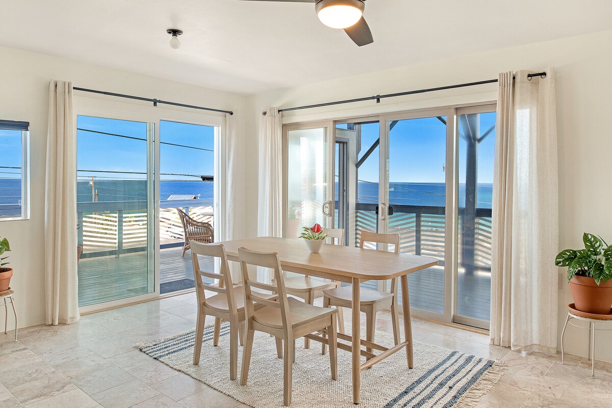 A light-filled dining area features a wooden table surrounded by four chairs, with large glass doors opening to a scenic ocean view. Soft curtains frame the windows, and plants on the floor add a touch of greenery to the space.