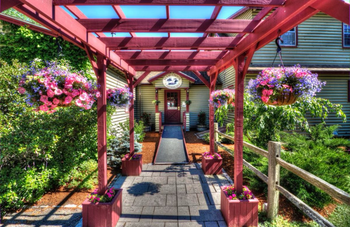 A red wooden arbor with hanging flower baskets frames the entrance to the inn. Colorful flowers bloom throughout the outdoor space, while a stone pathway leads visitors to the front door, surrounded by lush greenery and a wooden fence.