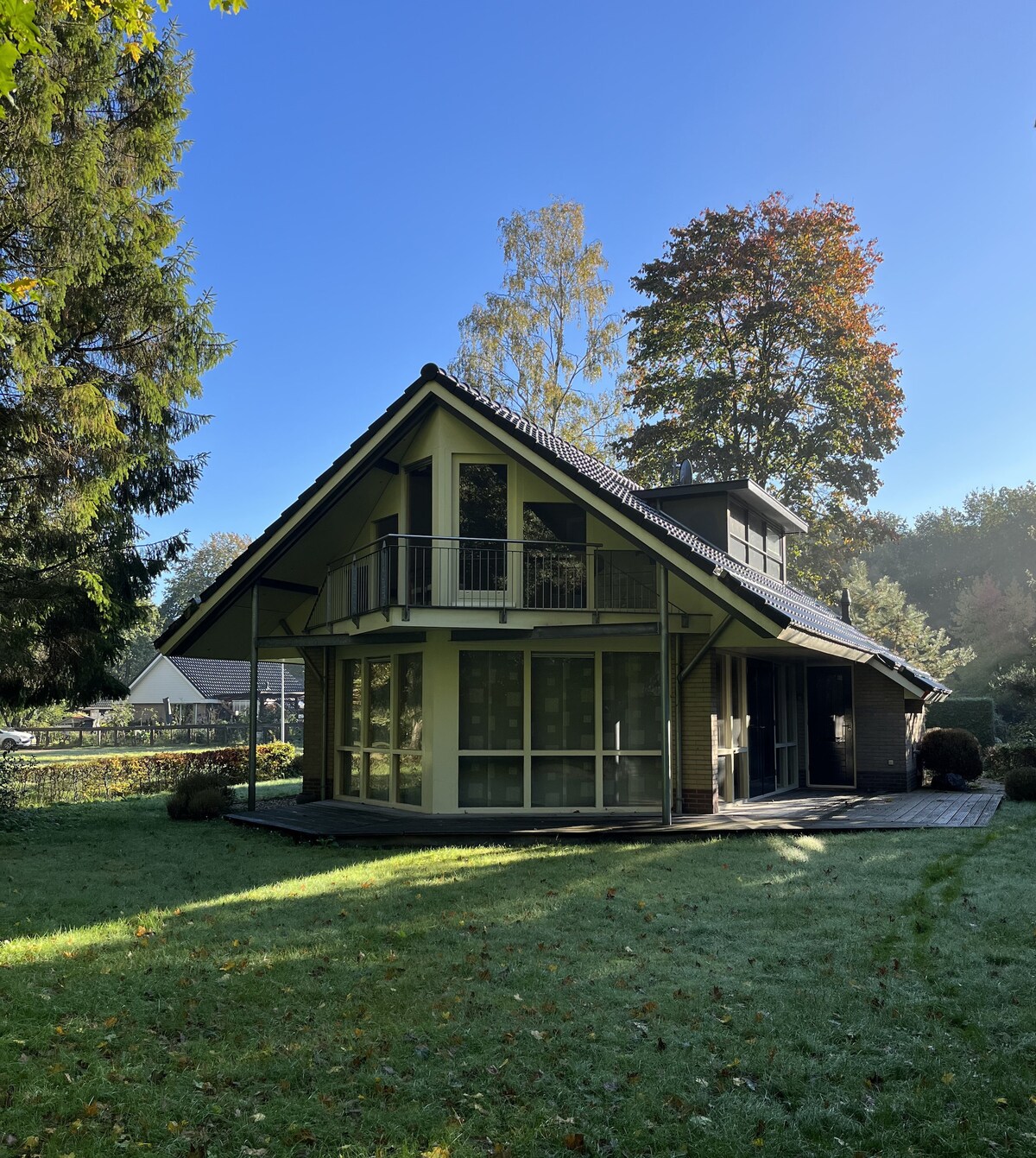 A charming house with angled roofs and large windows is surrounded by a lush green garden. The exterior features a combination of wood and stone, and a spacious deck extends outward. Trees and seasonal foliage create a serene backdrop under a clear blue sky.