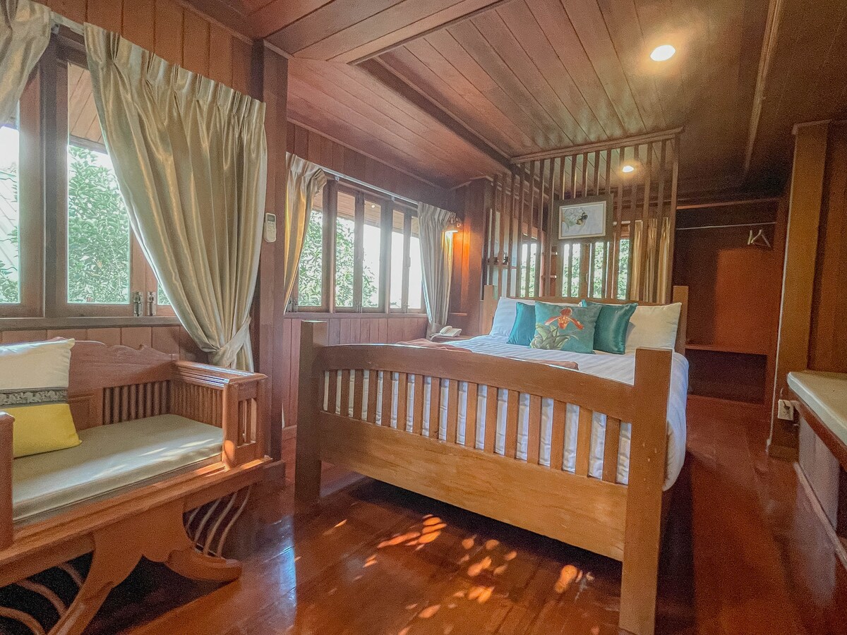 A wooden interior showcases a spacious sleeping area featuring large windows with soft drapes. A comfortable bed is complemented by decorative pillows, while a small seating area with intricate woodwork is present beside the bed. Natural light highlights the room's warm tones.