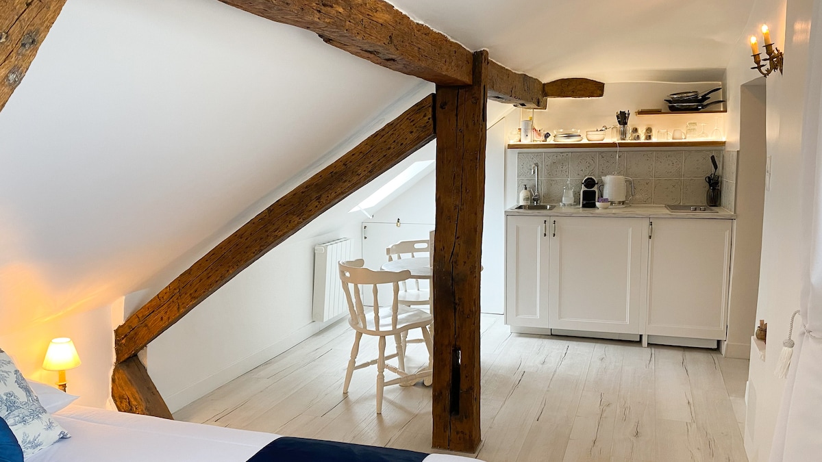 The studio features exposed wooden beams, creating a warm atmosphere. A cozy sleeping area is complemented by a well-equipped kitchenette, which includes essential appliances. A small dining table with two chairs is positioned nearby, allowing for comfortable dining in this compact space.
