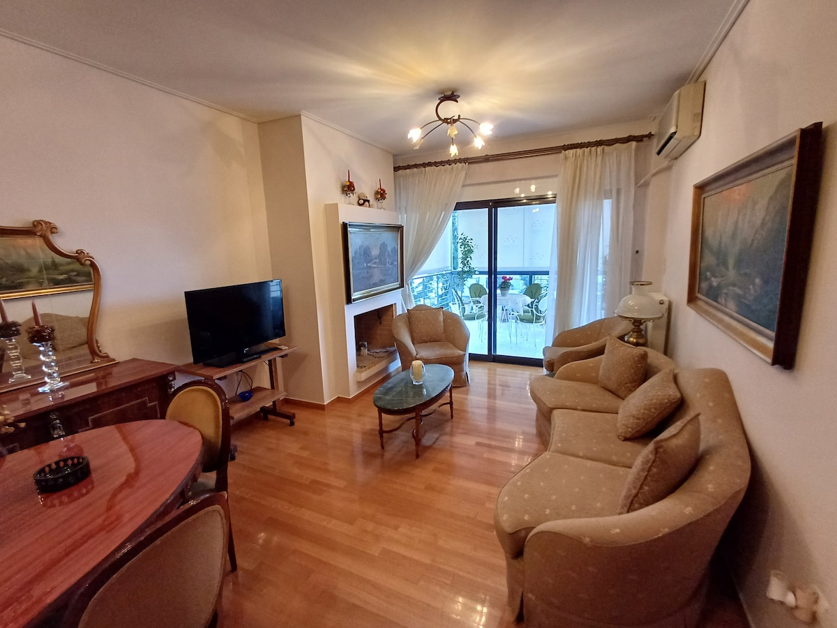 A spacious living area features a combination of plush seating and elegant furnishings. A wooden table is positioned near a television, and large glass doors open to a balcony, allowing natural light to fill the room. Soft curtains frame the view.
