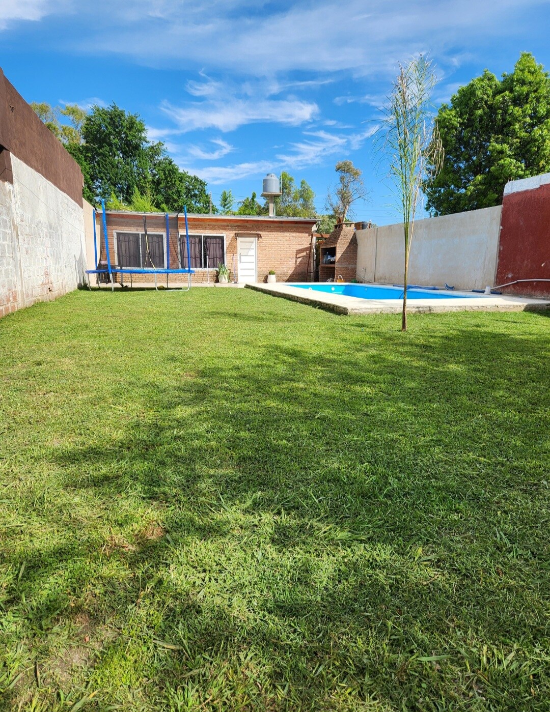 A large backyard is shown, featuring a grassy area and a swimming pool. A trampoline is visible near the house, with trees providing shade in the background. The sky is bright with scattered clouds, creating a peaceful outdoor environment.