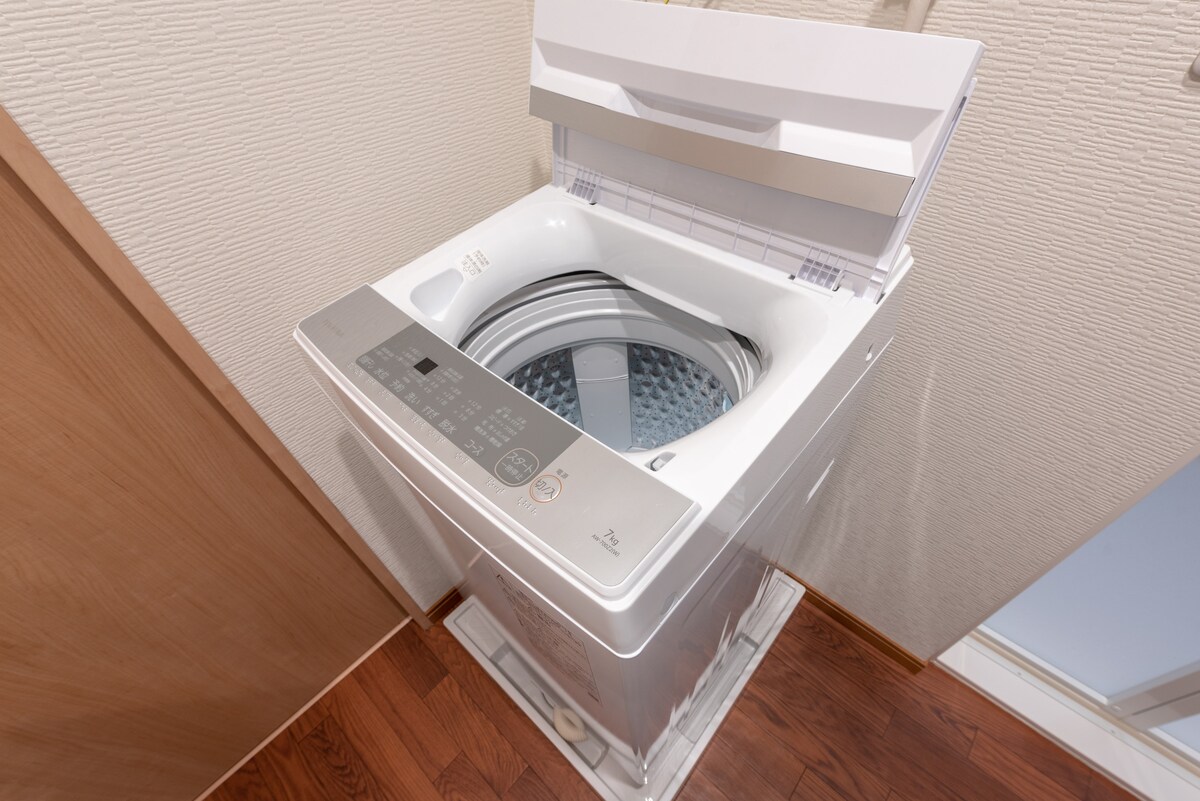 A modern washing machine is shown with the lid open, revealing a stainless steel drum. The control panel is displayed on the top front, featuring various buttons for different wash settings.