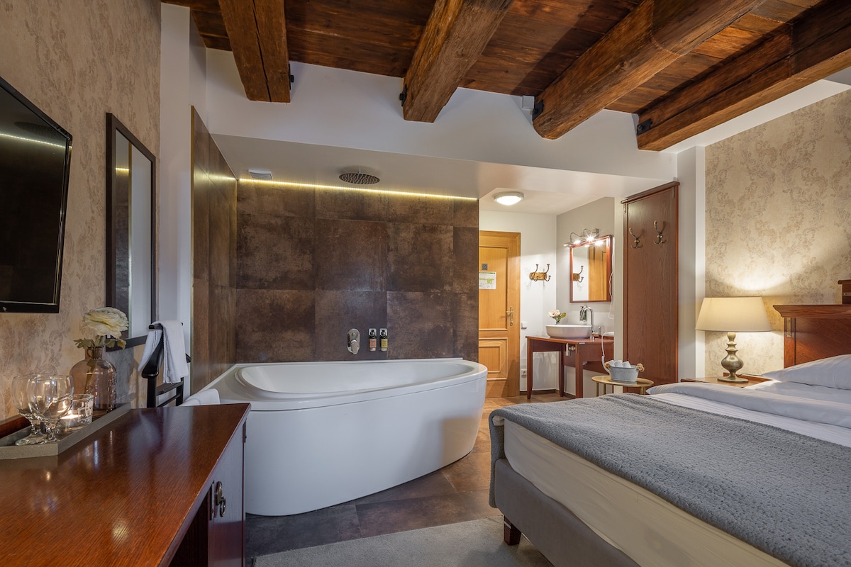 A spacious hotel room is highlighted by exposed wooden beams and a modern bathtub. A snug sleeping area is furnished with a comfortable bed, while a wooden desk and a mirror are visible across the room. Soft lighting enhances the relaxing ambiance.