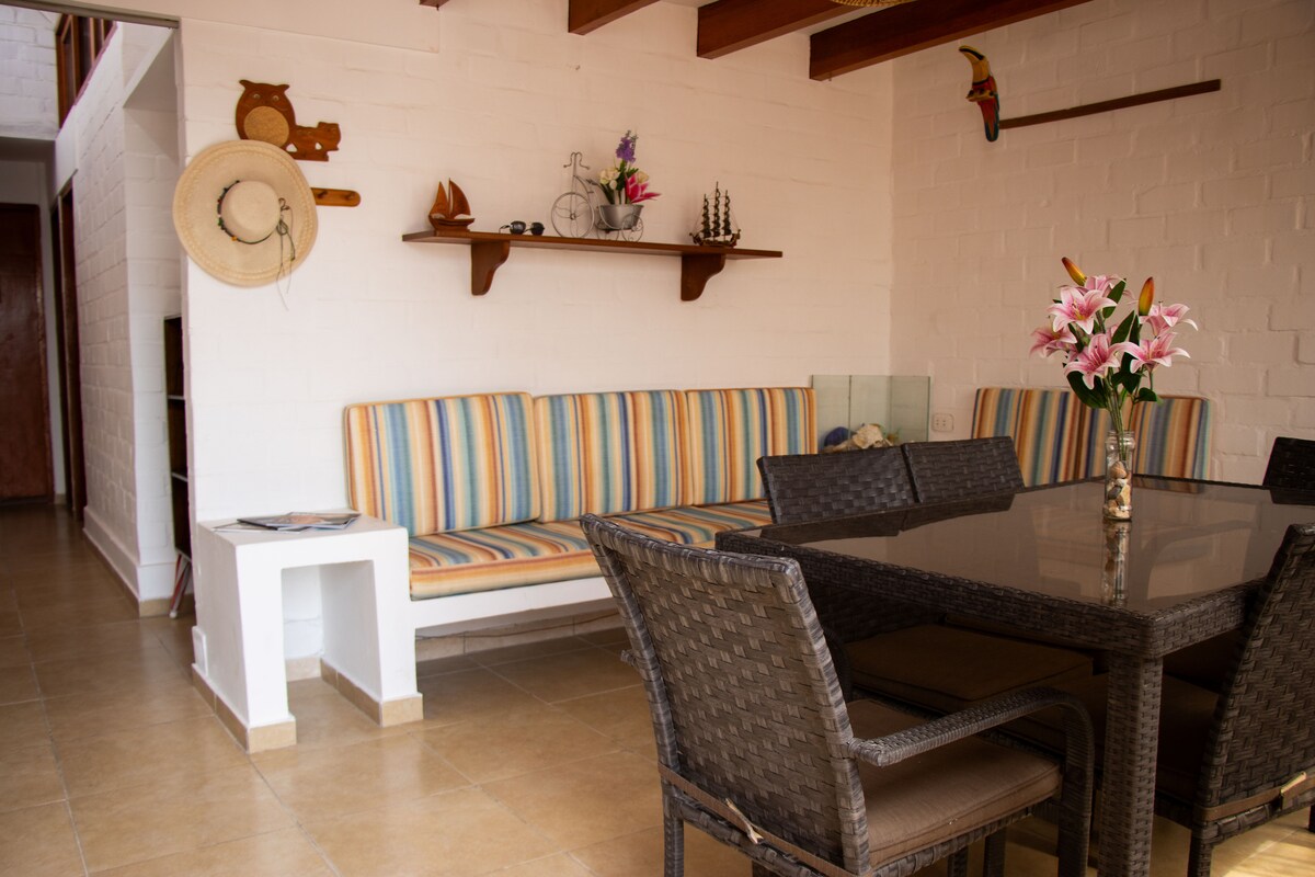 A cozy living area is presented with colorful striped seating against a white brick wall. A dark, glass-top dining table is surrounded by woven chairs. Decorative items and plants are displayed on a wooden shelf, enhancing the space's inviting atmosphere.