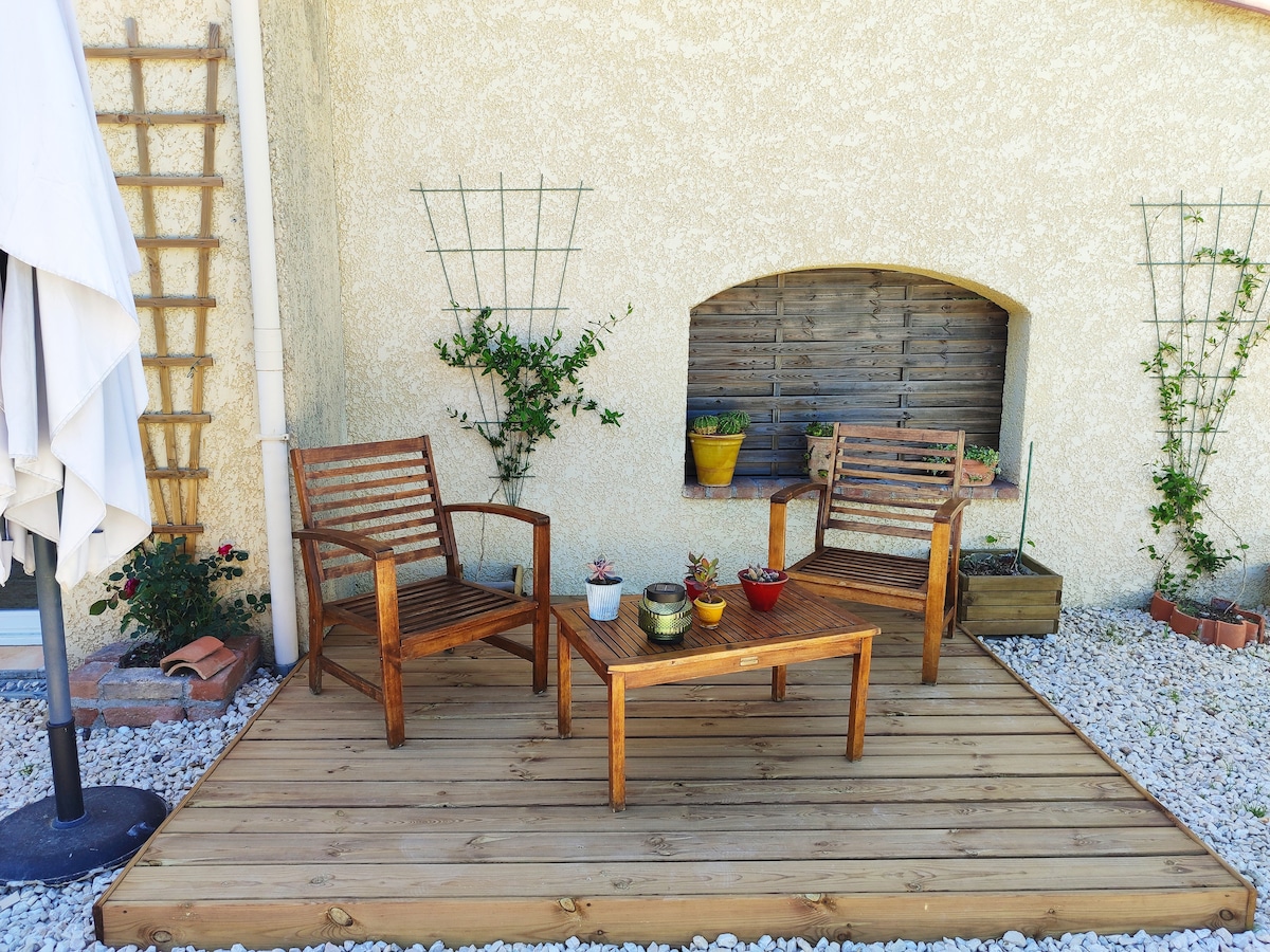 A small wooden deck features two slatted chairs positioned around a matching table. Potted plants are arranged on the ground and on a wall shelf, while climbing plants are supported by trellises. The area is surrounded by decorative gravel and a parasol is partially visible.