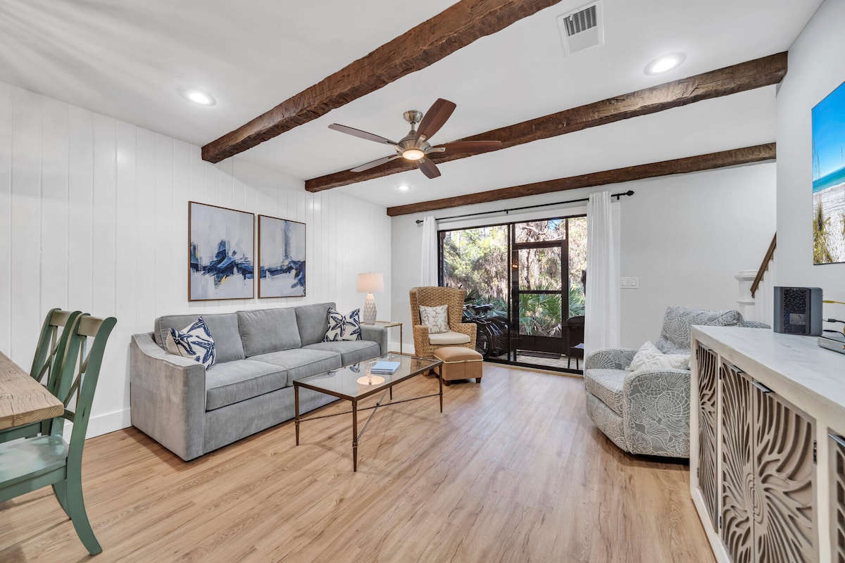The living area features wooden beams and warm wooden flooring. A cozy grey sofa and matching armchairs surround a coffee table. The space is illuminated by natural light from sliding glass doors leading to a private screened-in porch, enhancing the connection to nature.