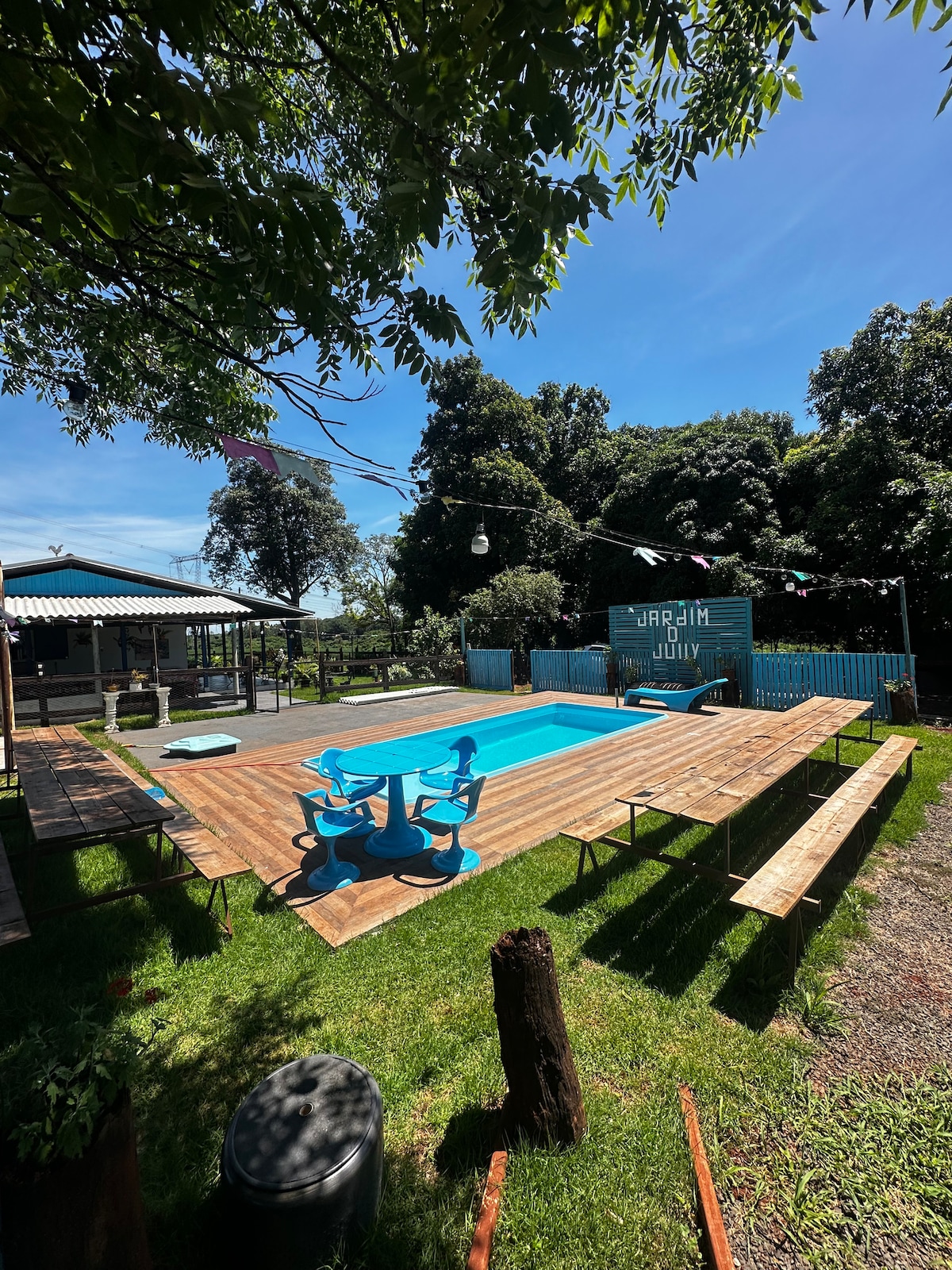 An outdoor area features a rectangular pool surrounded by wooden decking. Colorful picnic tables with blue seating are positioned nearby, set against a backdrop of lush greenery. A rustic building is visible in the background, enhancing the relaxed setting.