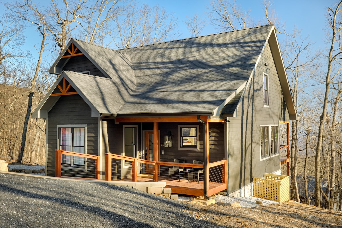 The exterior of Onyx Hill is composed of dark wood accents with a pitched roof, featuring large windows that allow for natural light. A welcoming porch extends to the front, furnished with several seating options, surrounded by trees and a gravel driveway leading up to the entrance.