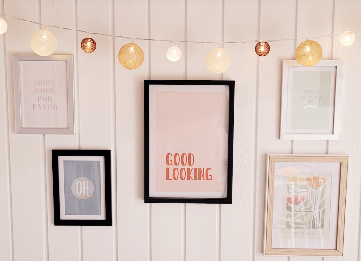 A selection of framed artworks adorns a wall, showcasing various styles and colors. A string of decorative lights adds a soft glow across the display, enhancing the visual appeal of the arranged pieces.