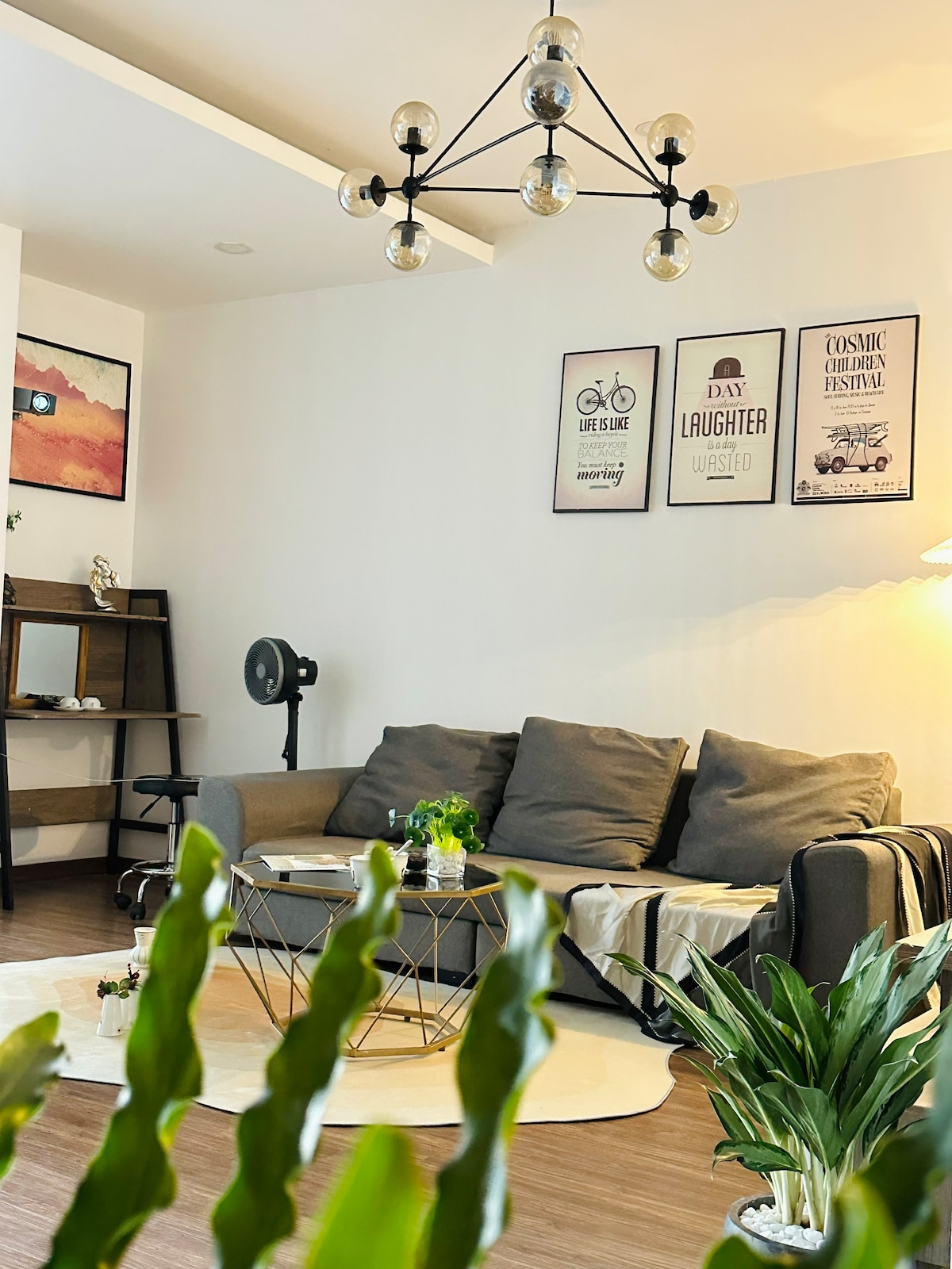 A living area is showcased with a spacious gray sofa and a stylish coffee table in the center. Light artwork adorns the walls, while a floor lamp and fan add functionality. Green plants are placed in view, contributing to the ambiance.