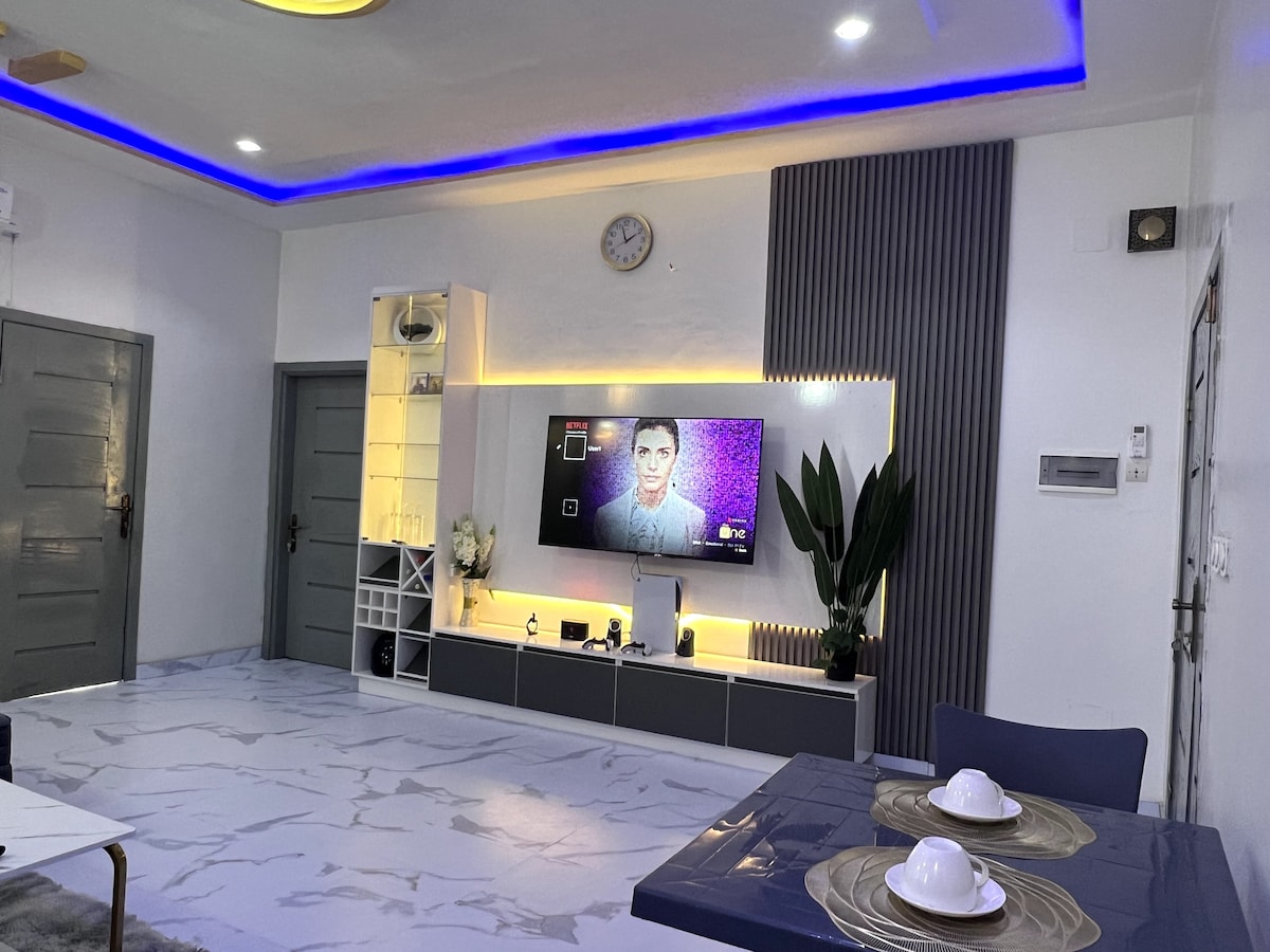 The living area features a modern television mounted on a stylish accent wall, illuminated by soft lighting. A sleek coffee table sits on a marbled floor, accompanied by a dining setup for two. Decorative plants add a touch of greenery to the space.