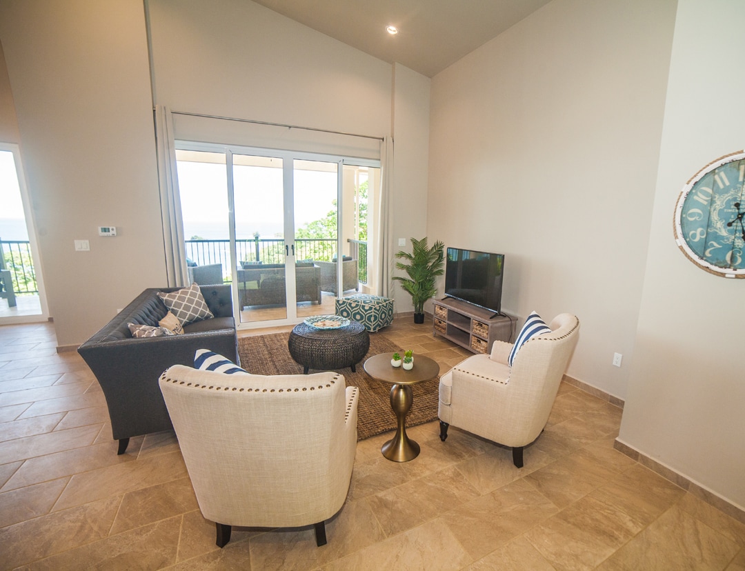 A spacious living area is presented with comfortable seating arranged around a central coffee table. Large glass doors lead to a balcony, allowing for natural light to fill the space and offering views of the ocean. A modern TV is positioned for easy viewing.