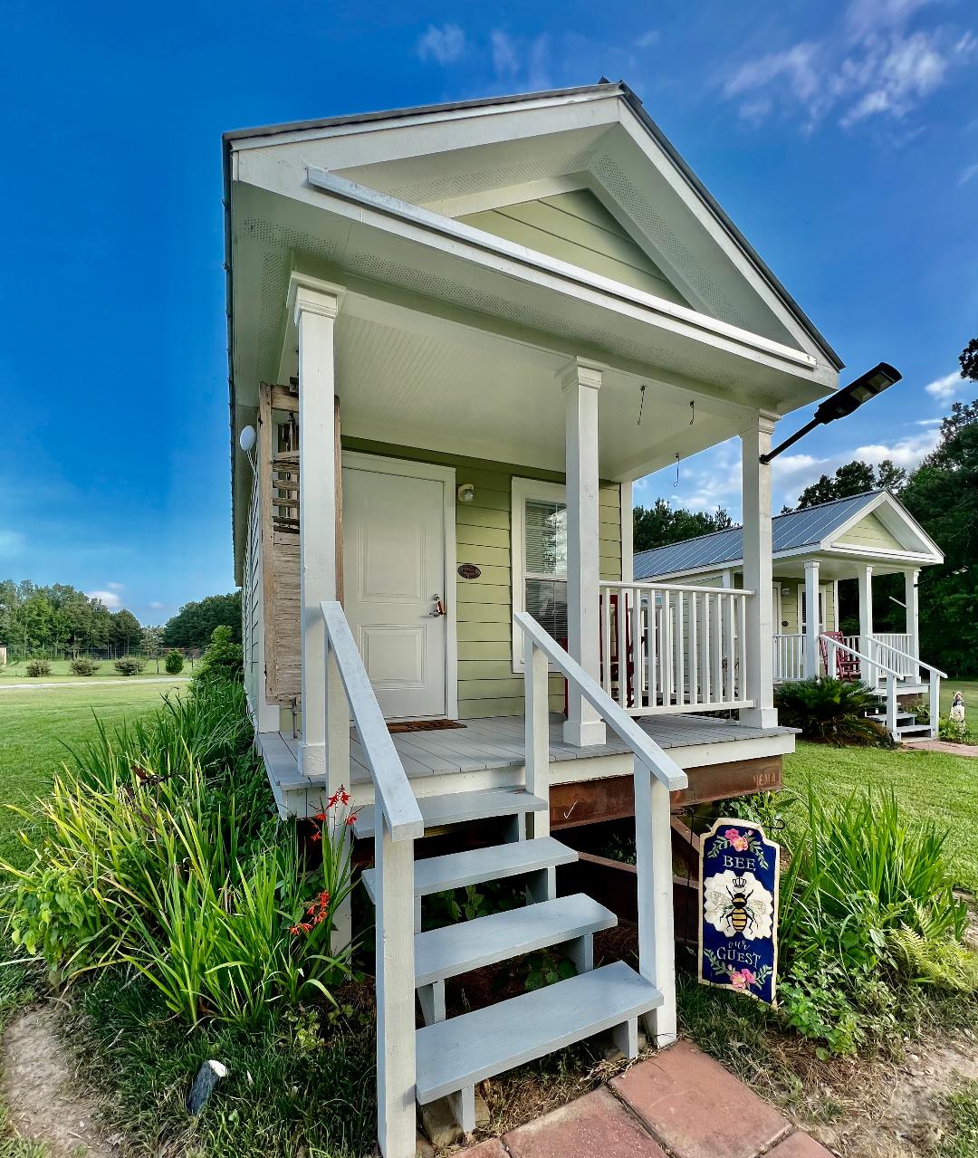 The exterior of a light green cottage is shown, featuring a gabled roof and white columns supporting the porch. Steps lead up to the front door, flanked by well-maintained greenery. A decorative sign stands nearby, indicating the cottage's name.