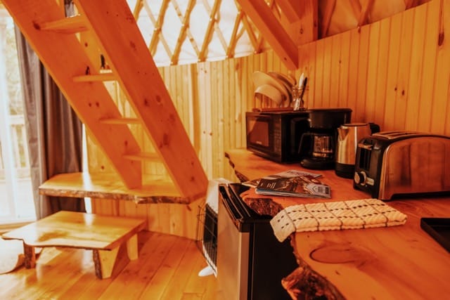 The interior of the yurt features a warm wooden design with a kitchenette area. A small dining table with a bench is visible beside the counter, which includes a microwave, coffee maker, and kettle. A staircase leads to the lofted bedroom, and natural light filters through nearby windows.