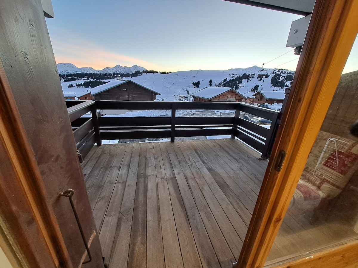 A wooden balcony is shown, featuring a spacious deck with wooden flooring. The view overlooks snow-covered mountains and a serene landscape during sunset, with scattered wooden chalets in the foreground, conveying a sense of tranquility.