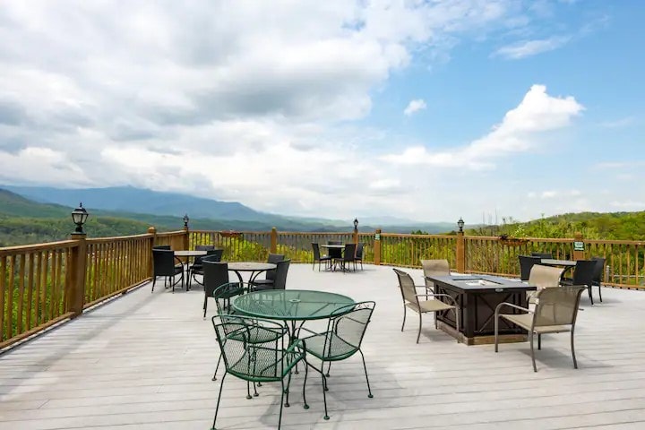 An outdoor deck features multiple seating arrangements, including tables with glass tops and metal chairs. The expansive space offers sweeping views of the surrounding mountains under a partly cloudy sky, providing a serene setting for relaxation.