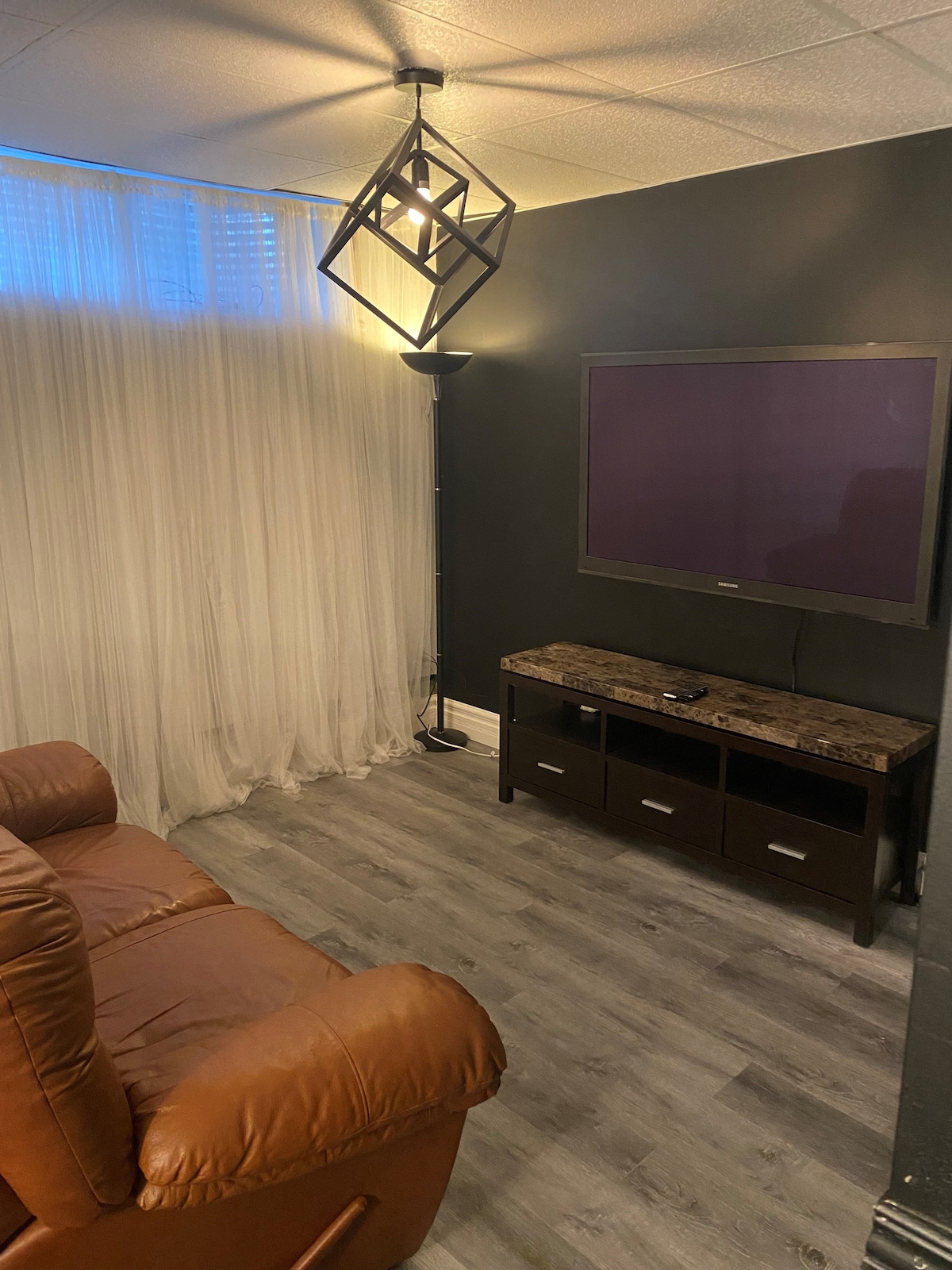 A comfortable seating area features a brown recliner positioned near a large wall-mounted television. The space includes a dark, low-profile media console with multiple drawers. Soft drapery allows natural light to filter in, and a modern light fixture provides additional illumination.