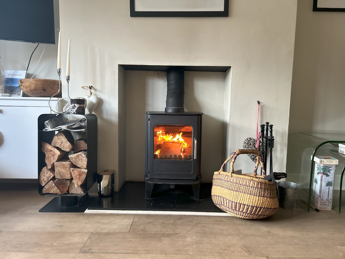 A cozy wood-burning stove is situated in a minimalist fireplace, radiating warmth with a visible flame. Firewood is neatly stacked beside it, and a woven basket lies nearby. Various tools for tending the fire are displayed, complementing the inviting atmosphere of the living area.
