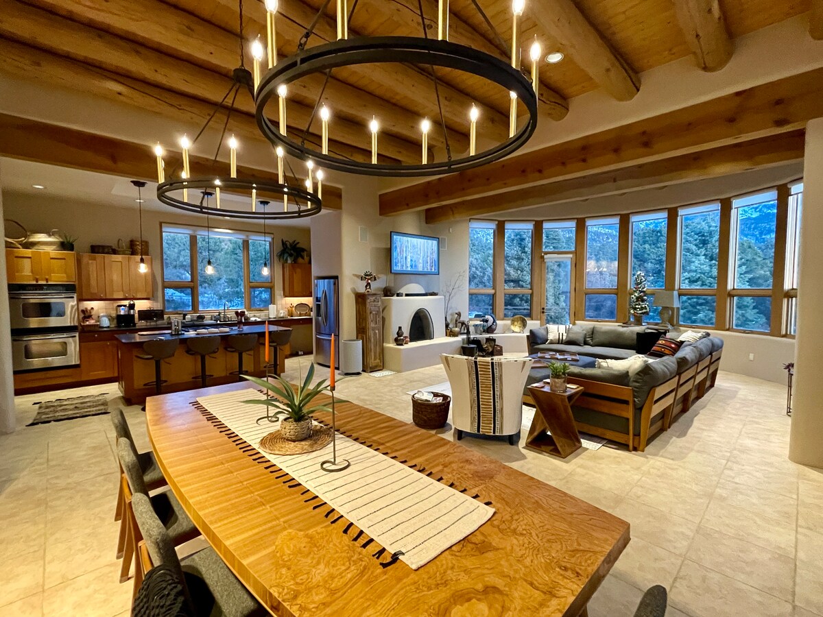 An open-concept living area features wooden beams and large windows that provide sweeping views of the surrounding landscape. A long wooden dining table is paired with dark chairs, and a spacious sectional sofa is positioned in the relaxation area, beneath modern light fixtures.