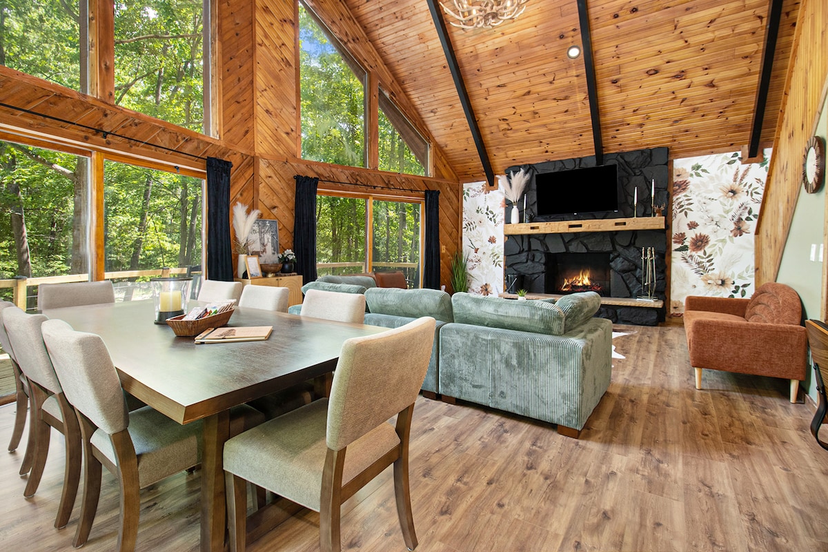 A spacious living area is framed by large windows that invite natural light, showcasing a mix of wood and stone textures. Comfortable seating includes a sofa and armchair, arranged around a central coffee table. A dining table for eight is positioned nearby, enhancing the gathering space.