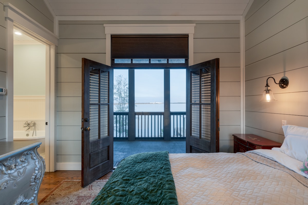 Large, open doors reveal a serene lake view from a cozy bedroom. The inviting bed features a soft quilt, while a vintage dresser adds character to the space. Light enters through the doors, illuminating the calming interior with painted walls.