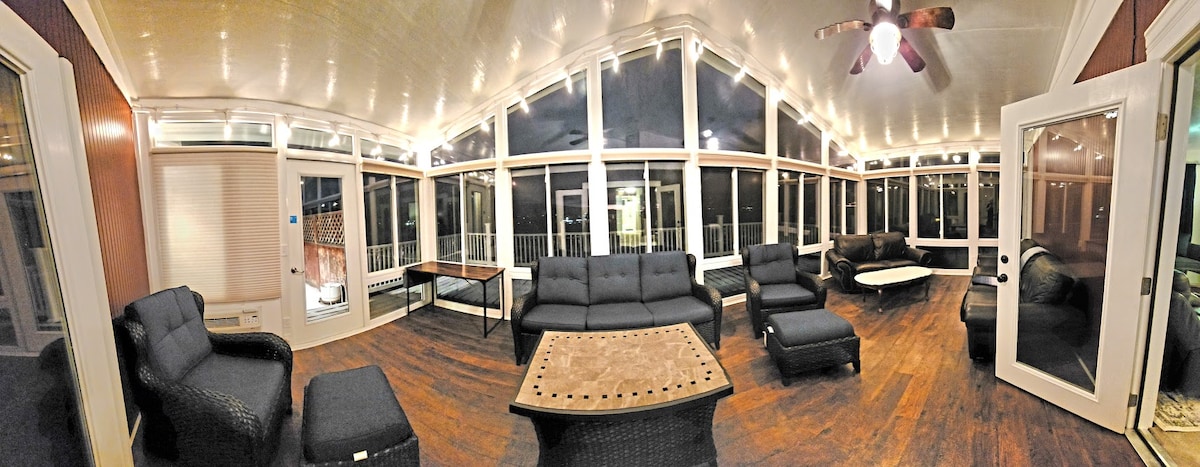 A spacious sunroom features large windows that provide ample natural light. Comfortable seating is arranged around a central coffee table, with dark wicker furniture complementing the warm wooden flooring. Ceiling fans and overhead lighting enhance the open atmosphere.