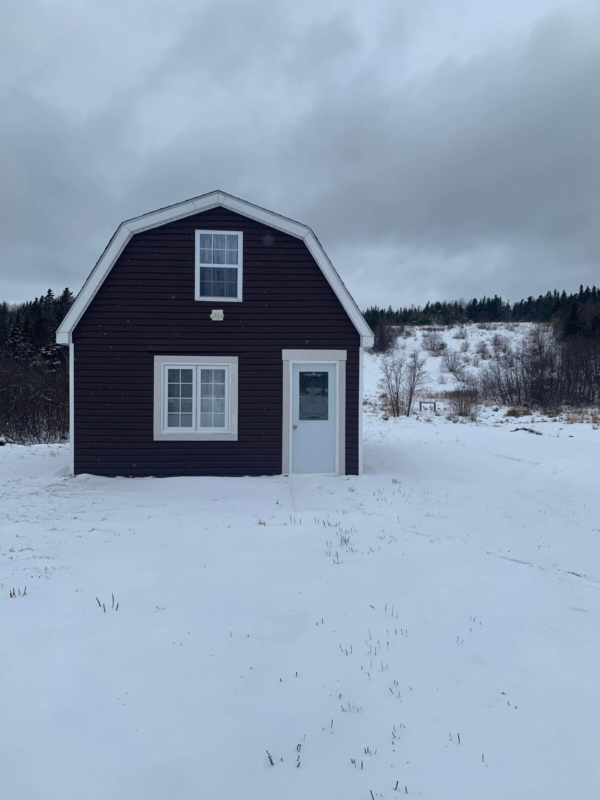 A quaint, two-story cabin features a classic A-frame roof and is set against a snowy landscape. A front door and two windows allow natural light to enter, while the surrounding area showcases gentle slopes of white snow and scattered trees in the background.