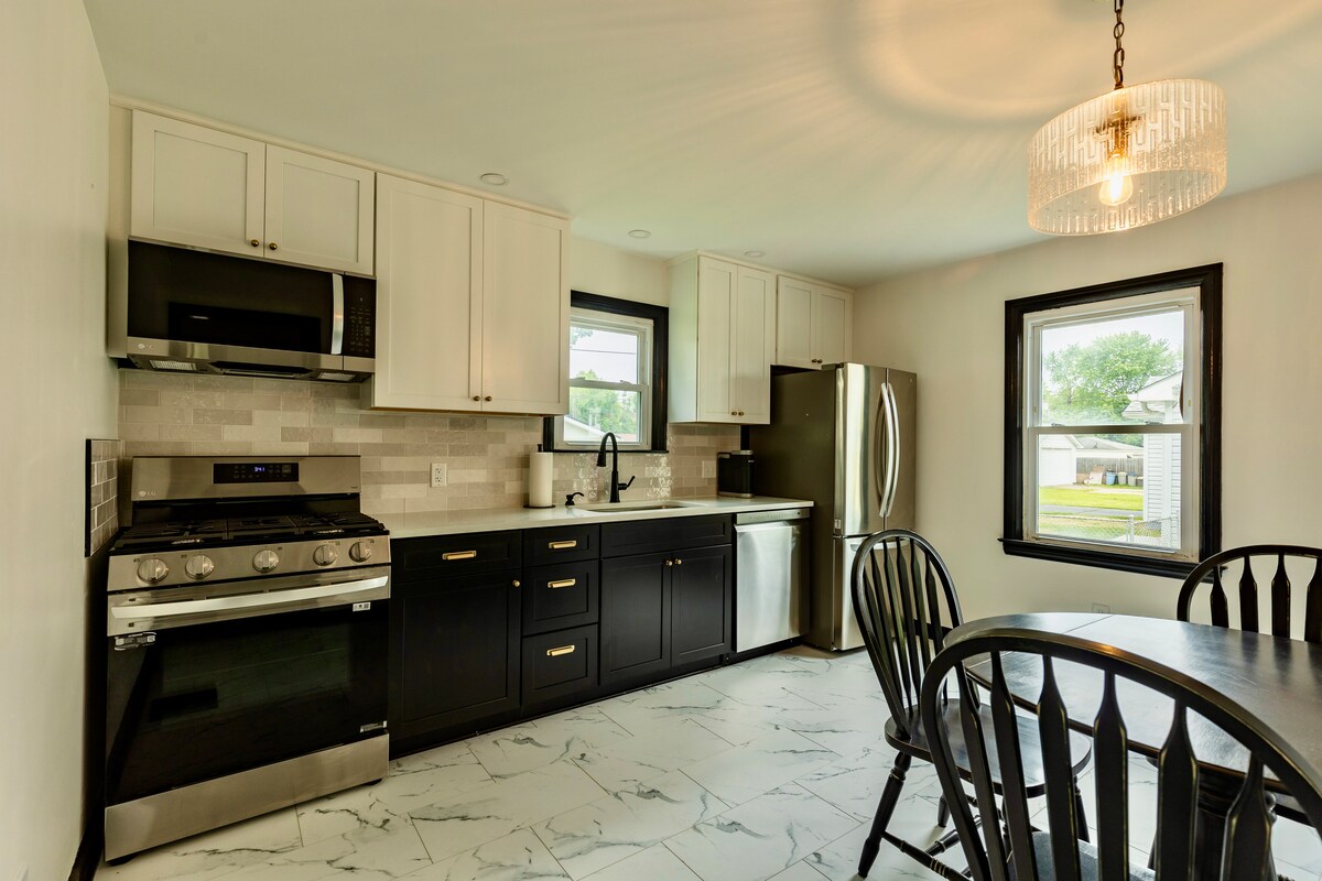 A modern kitchen features sleek black cabinetry and stainless steel appliances. A gas stove and oven sit beneath a microwave, while a dishwasher is positioned beside a spacious refrigerator. A dining table with black chairs occupies one corner, illuminated by a stylish overhead light.