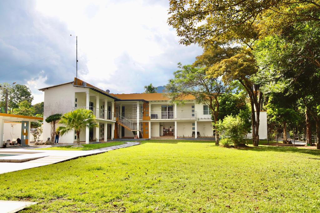 The exterior view of a two-story house is surrounded by lush greenery and several mature trees. A spacious lawn in front invites relaxation, while large windows reflect natural light. A pathway leads towards the entrance, showcasing a welcoming and open layout.