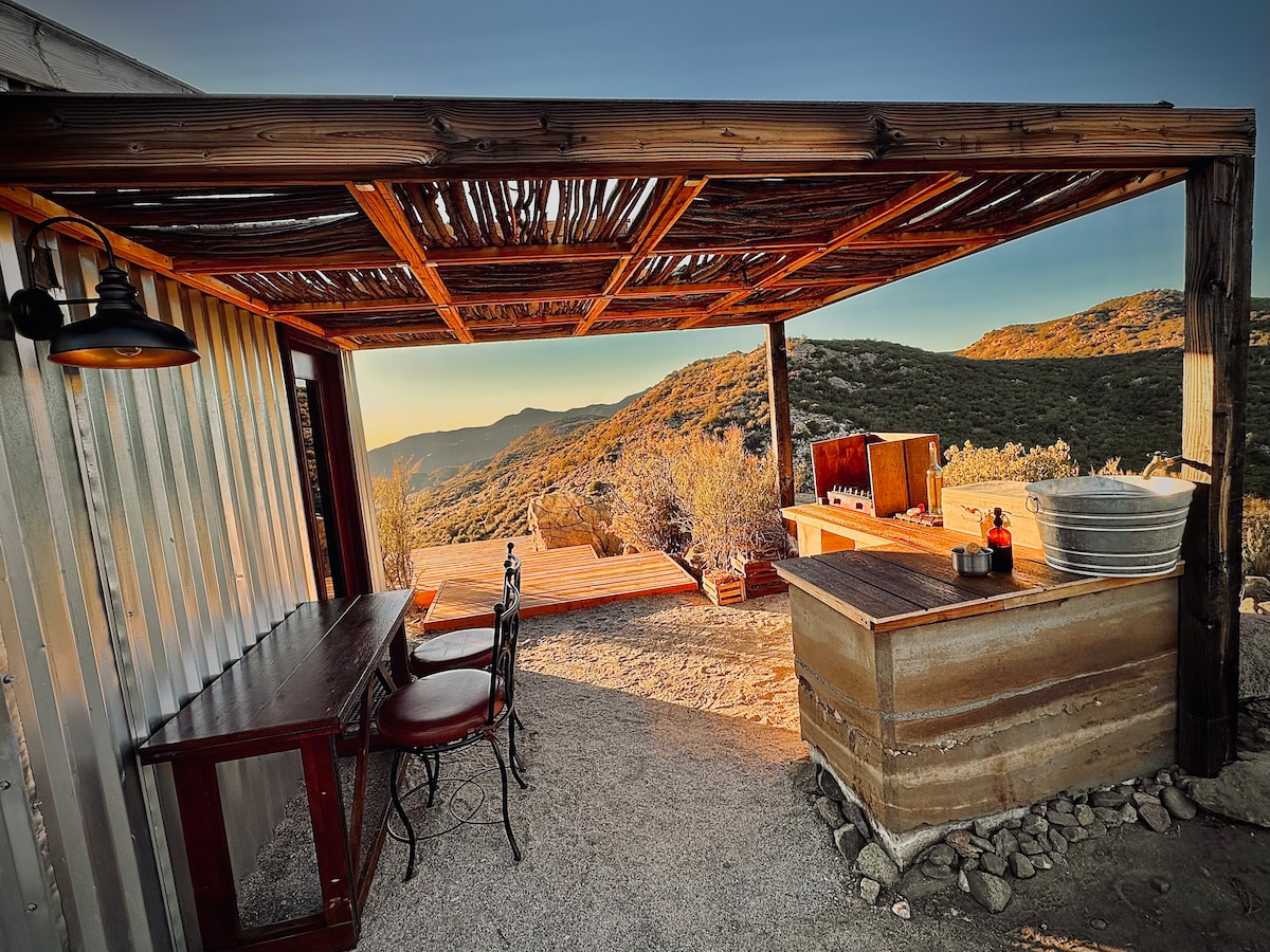 An outdoor cooking and dining area is framed by a wooden pergola, with a rustic table and two chairs positioned nearby. A barbecue grill is located next to a large, metal wash basin. The surrounding landscape features rolling hills and a colorful sunset in the background.