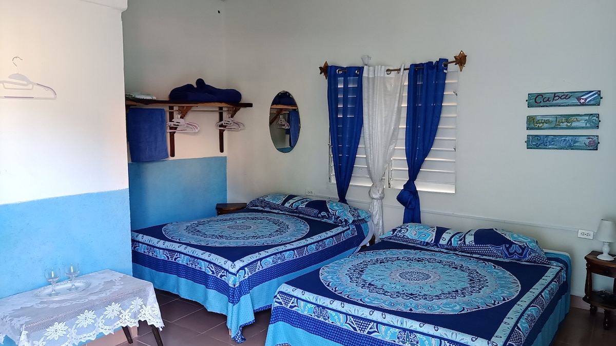 A bright room features two spacious beds, each adorned with blue-patterned bedding. A mirror reflects light from a window with white curtains. Shelves hold blue towels, while a small table with two glasses adds a touch of convenience to the space.