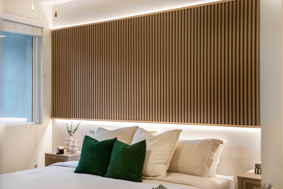 A serene bedroom features a king bed adorned with soft linens and two decorative pillows. A textured wood panel serves as a headboard, softly illuminated by recessed lighting. A nightstand with decorative items is positioned on either side, while natural light filters through a nearby window.