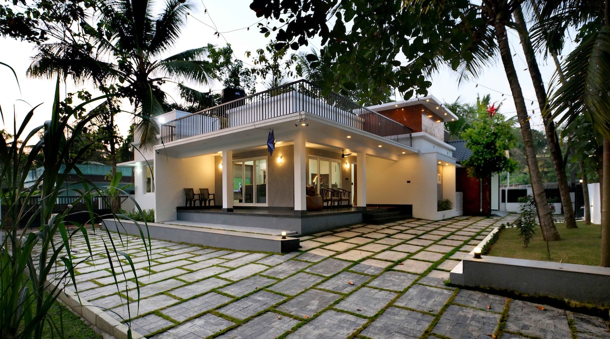 The exterior of the villa is characterized by a modern design, featuring spacious terraces and a large patio surrounded by neatly arranged stone paving. Lush greenery and palm trees provide a natural backdrop, enhancing the tranquil ambiance of the property.