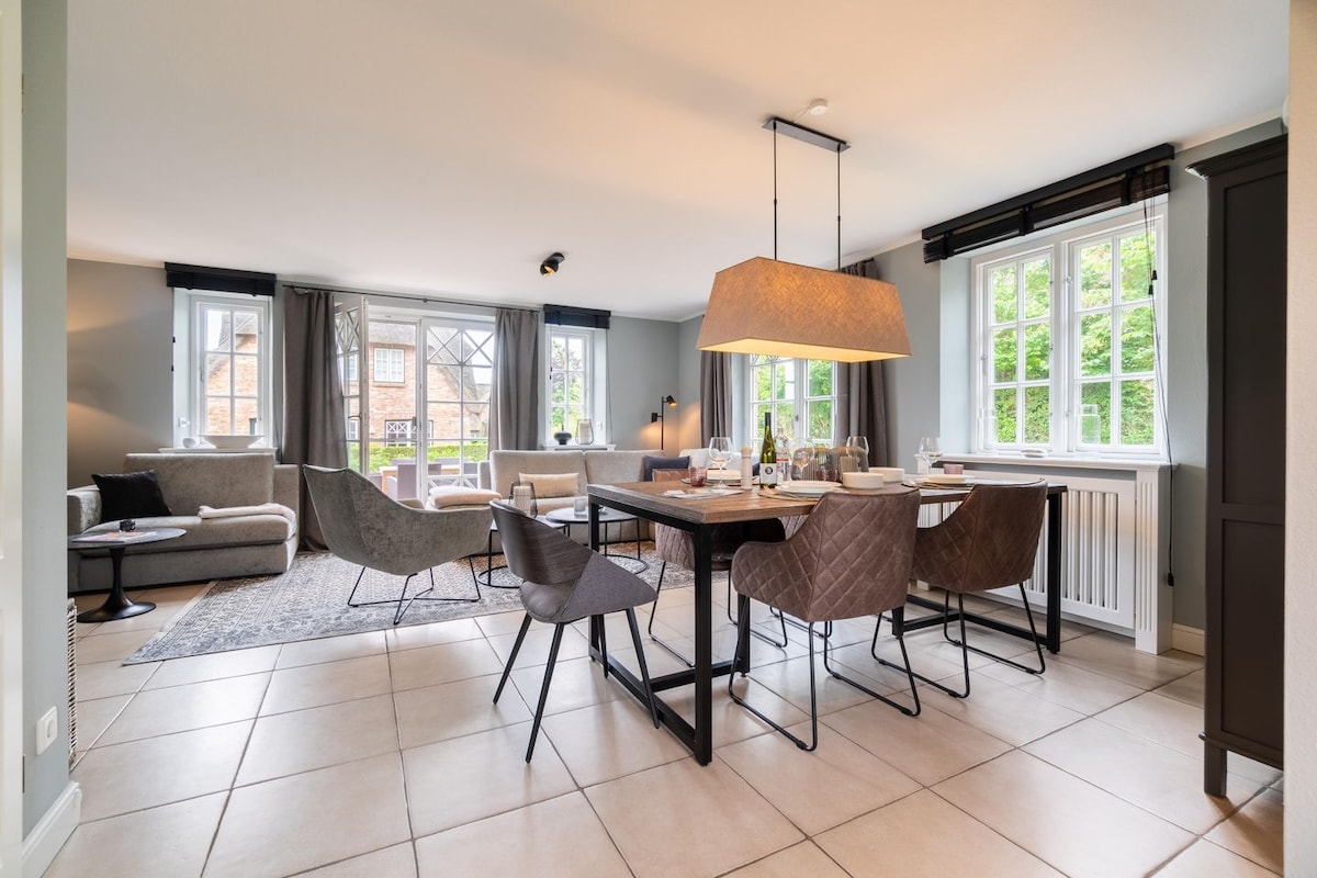 An open living area features a dining table with elegant chairs positioned near large windows, allowing natural light to illuminate the space. A comfortable seating arrangement is visible, set against a neutral palette and minimal decor, creating a calm environment.