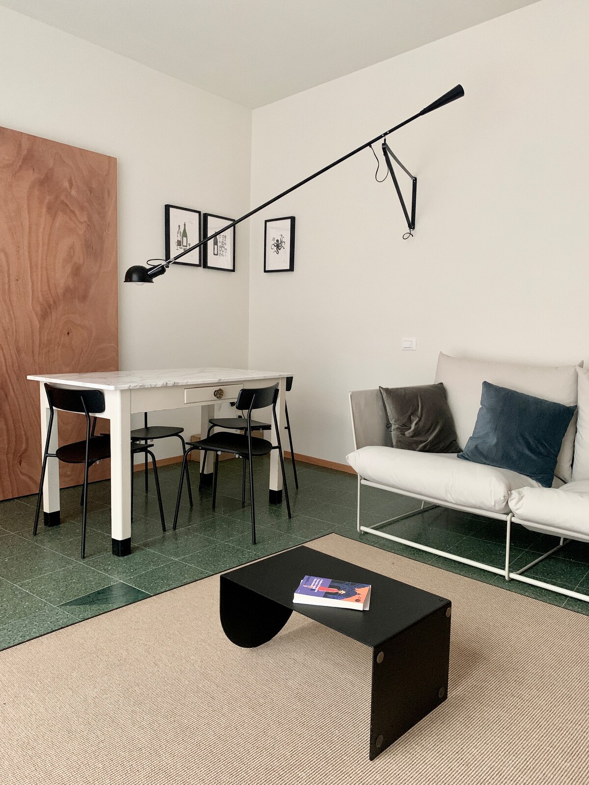 A modern living space is depicted with minimalist furniture. A sleek white table is paired with black chairs. A light fixture hangs from the ceiling above the table. A small black coffee table sits in front of a comfortable light sofa adorned with blue cushions.