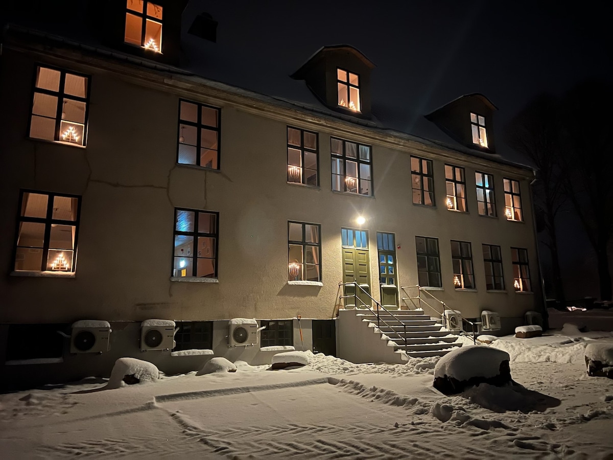 The exterior of a charming, two-story building is depicted at night, illuminated by lights in the windows. Snow covers the ground and stairway, creating a serene winter scene. Air conditioning units are affixed to the lower walls, and steps lead to a prominent green door.