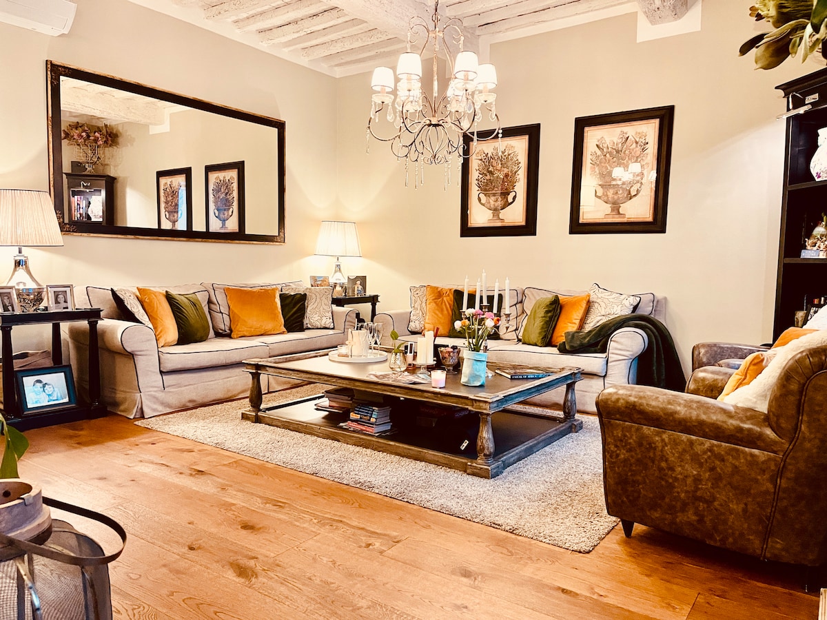 A cozy living room is presented with soft beige walls and wooden flooring. Two comfortable sofas with decorative cushions face a wooden coffee table. Wall art with floral themes and a large mirror are displayed. A chandelier provides soft lighting, enhancing the inviting atmosphere.