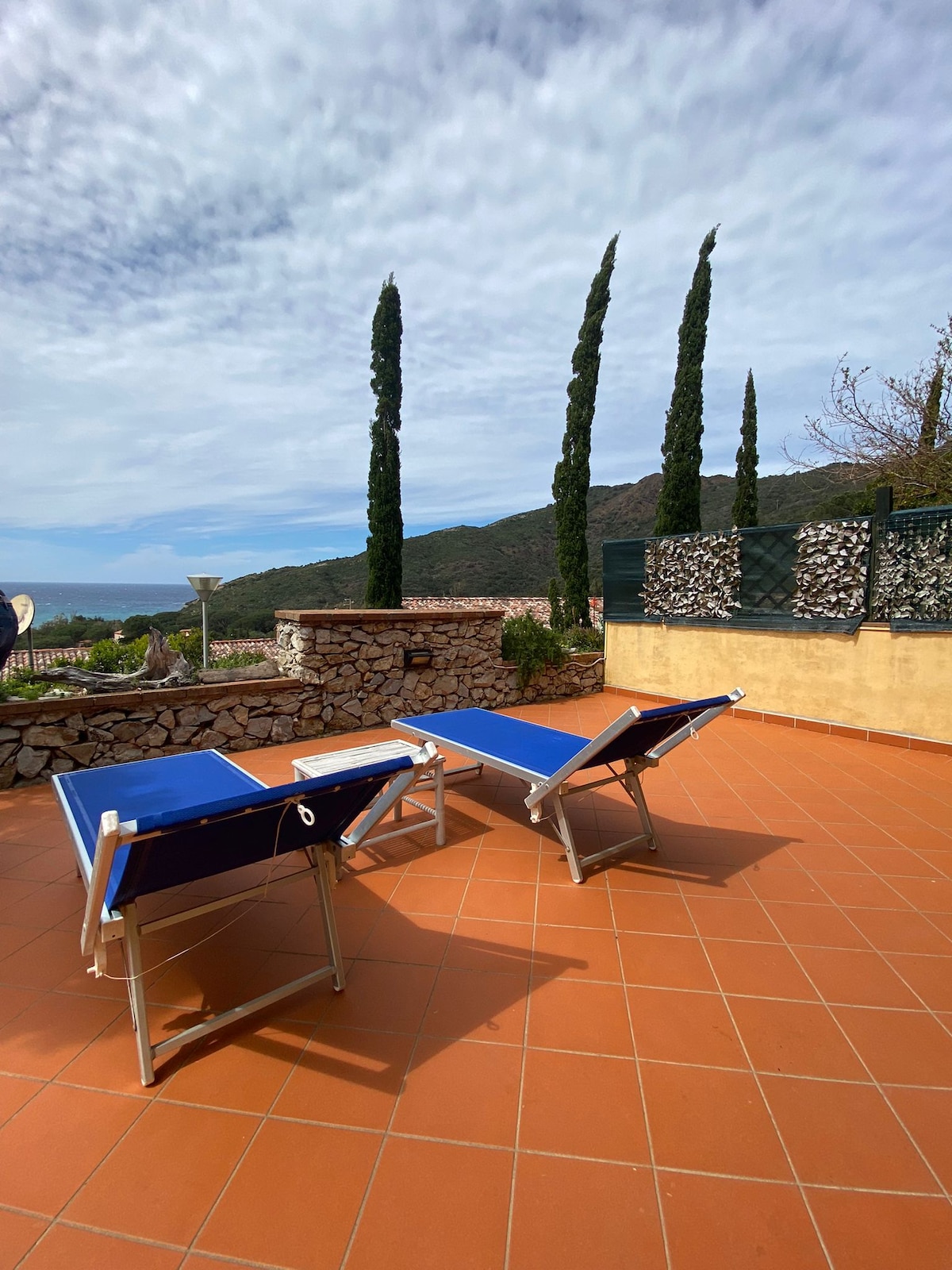 Two blue lounge chairs are positioned on a spacious tiled terrace, offering a serene view of the surrounding hills. Tall, slender evergreen trees are visible in the background, surrounded by a stone wall, contributing to the peaceful outdoor setting.