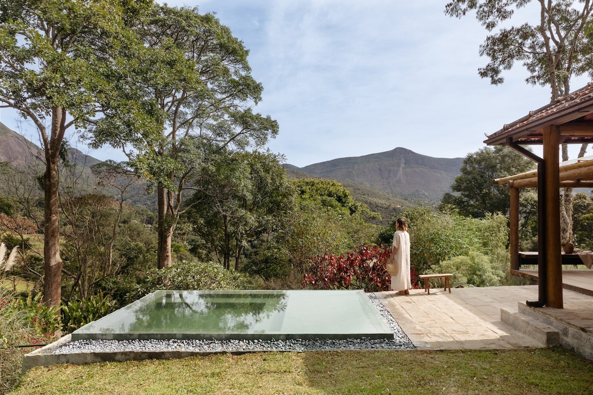 An outdoor area features a clear swimming pool surrounded by natural greenery. A wooden structure is visible to the right, and a person is seen sitting peacefully on a bench, with mountains providing a serene backdrop.