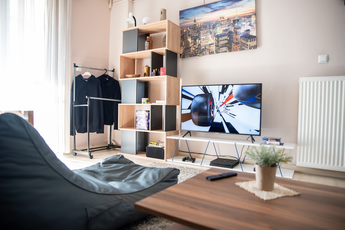 A modern living area showcases a large flat-screen television against a stylish shelving unit filled with decorative items. A cozy gaming pouf is placed in front of a wooden coffee table, with a small plant adding a touch of greenery. Natural light floods the space through sheer curtains.