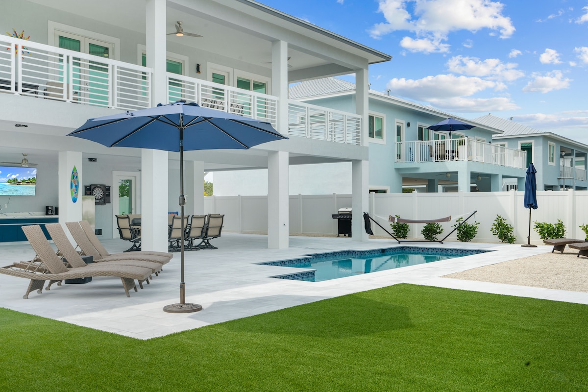 The outdoor area features a heated swimming pool surrounded by well-maintained grass. Sun loungers with neutral tones and an umbrella provide shaded seating. A dining table with multiple chairs is set under the elevated deck, which overlooks the space, ideal for relaxation and outdoor dining.