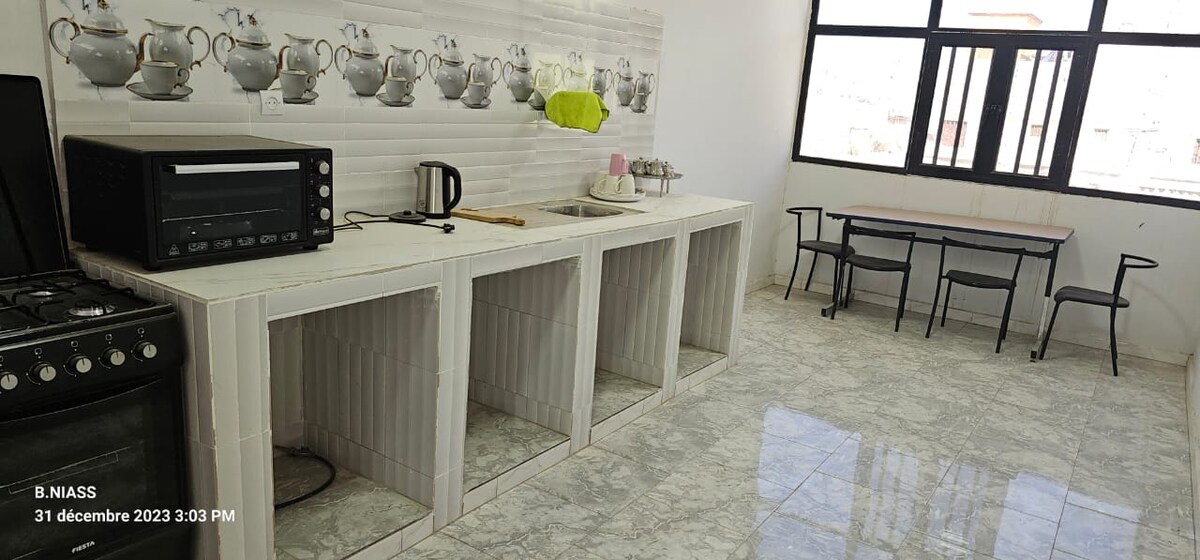 A bright kitchen is featured with a modern gas stove, an oven, and a kettle. The white cabinetry is complemented by tea sets arranged on the wall. A dining table with four chairs is positioned near the large window, allowing natural light to fill the space.