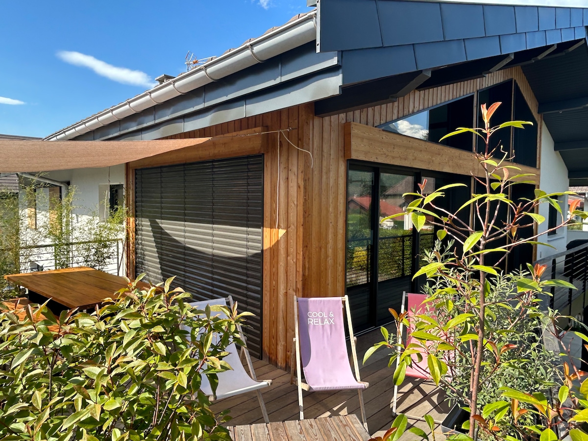 A wooden-clad terrace is visible, showcasing outdoor seating in pastel colors. Lush greenery surrounds the area, complemented by retractable awnings over large glass windows. Natural light enhances the inviting ambiance of the space.