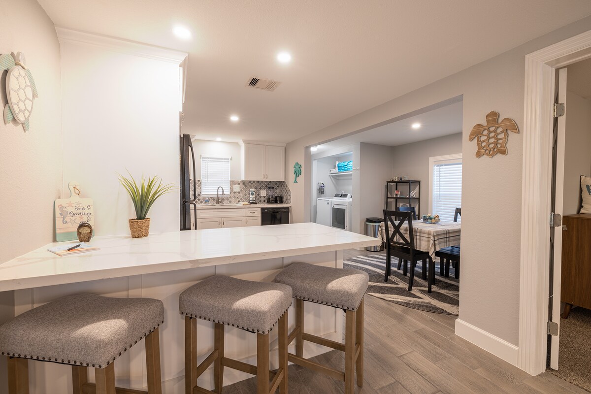 A modern kitchen is showcased, featuring sleek cabinetry and a spacious island with bar seating. Natural light brightens the area, illuminating the tiled backsplash and stainless steel appliances. An open layout connects the kitchen to a cozy dining space in the background.